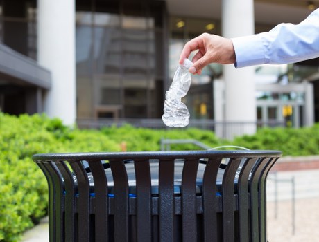 A person's hand is seen dropping a crumpled plastic water bottle into a black outdoor rubbish bin situated on a paved area. The hand appears to be wearing a light blue shirt or jacket. In the background, there is a modern building with large glass windows and a white exterior, along with some green bushes and red brick paving. The scene is outdoors, likely in an urban or commercial area within Newham, and the image emphasizes the act of disposing of recyclable waste properly. This visual supports waste management and rubbish removal services provided by Business Waste Removal Newham, highlighting responsible waste disposal practices in a local context.