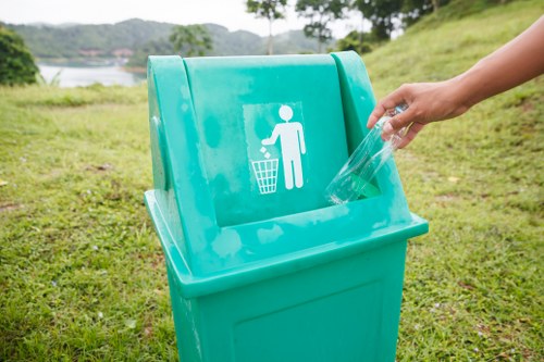 A close-up of a bright turquoise outdoor rubbish bin with a white pictogram of a person disposing of waste, situated on a grassy area near a body of water and trees in the background. A person’s hand is seen placing a clear plastic bottle into the bin, which is positioned on a lawn with natural grass and small patches of dirt. The scene appears to be in a park or riverside location, consistent with waste disposal and litter collection services in Newham. The bin has a smooth finish with a hinged lid, and the environment is illuminated with natural daylight, emphasizing the outdoor setting for rubbish collection and waste management activities.