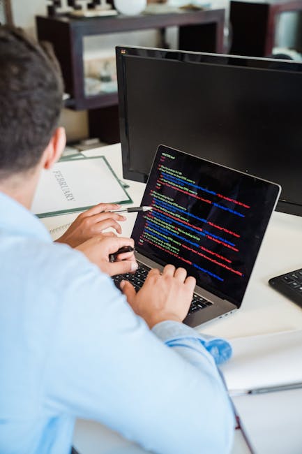 A person seated at a desk working on a laptop with a black screen displaying colorful lines of code, sitting in front of a second large monitor that is turned off. The individual has short dark hair a