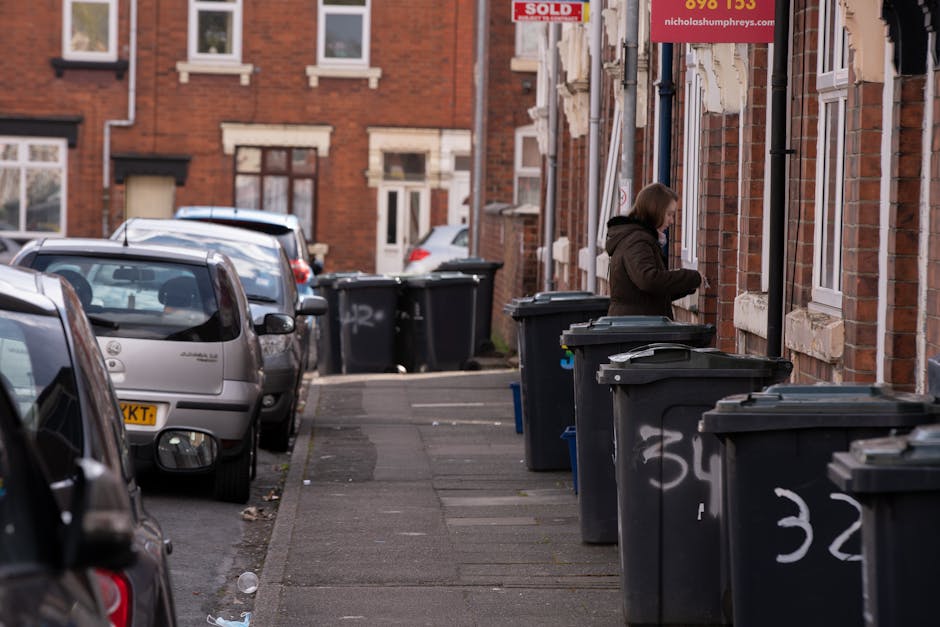 A middle-aged woman with short dark hair, dressed in a black t-shirt and dark trousers, is seen on a paved outdoor walkway or park area, bending slightly forward as she disposes of large white rubbish